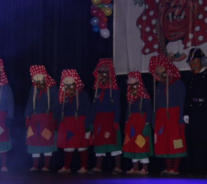 Fünf Personen in bunten Kostümen mit roten Kopftüchern mit weißen Punkten und Holzmasken stehen nebeneinander auf einer Bühne.