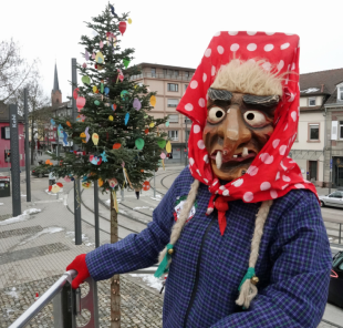 Person mit rot-weiß gepunktetem Kopftuch, blauer Jacke und roten Handschuhen steht vor einem geschmückten Baum auf einem Platz mit Straßenbahnschienen.