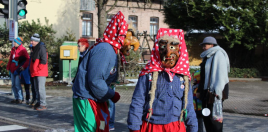 Person in historischer Uniform mit Säbel und Person in bunter Tracht mit rotem Kopftuch, Zöpfen und Holzschuhen vor rotem Fahrzeug mit Plakat auf dem Dach bei Straßenumzug.