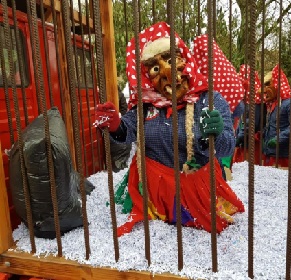 Figur mit Hexenmaske, rotem Kopftuch mit weißen Punkten, blauem Oberteil und rotem Rock steht hinter Eisenstäben auf weißem Streugut.